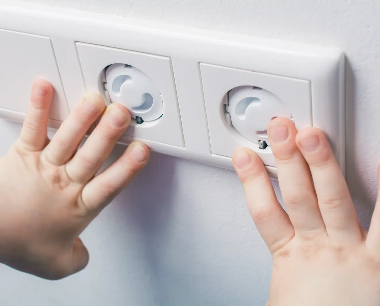 Close-up of a child’s hands touching a triple electrical outlet with built-in childproof safety covers on a white wall.