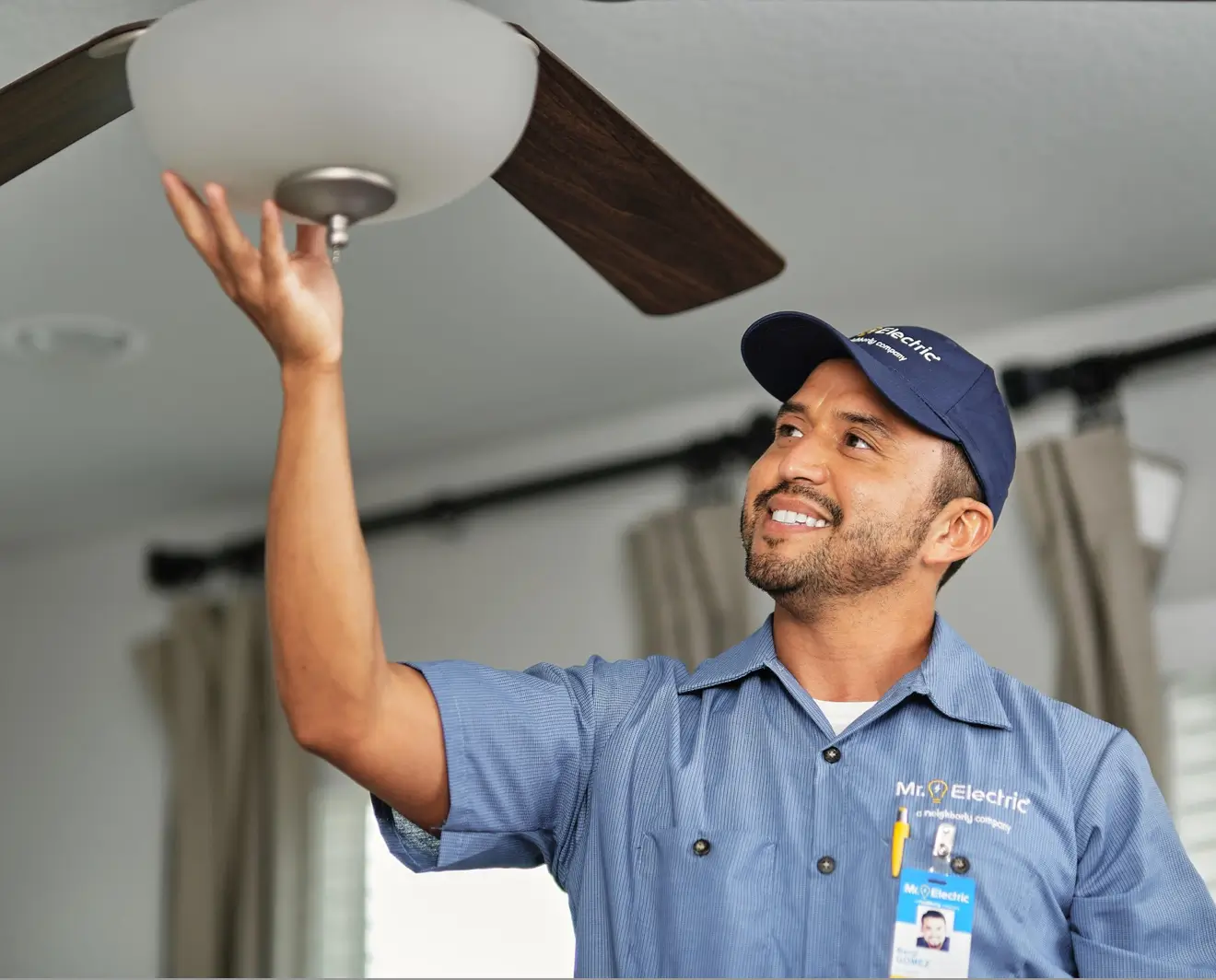 Smiling male electrician wearing a blue company uniform and cap, standing on a ladder as he adjusts a modern ceiling fan light indoors with beige curtains in the background.