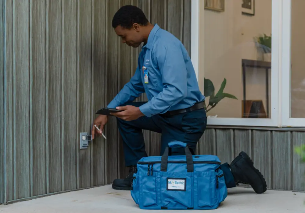 Male electrician in a blue uniform kneeling on a patio while inspecting an outdoor wall electrical outlet using a tool, with a blue tool bag beside him and a house window in the background.