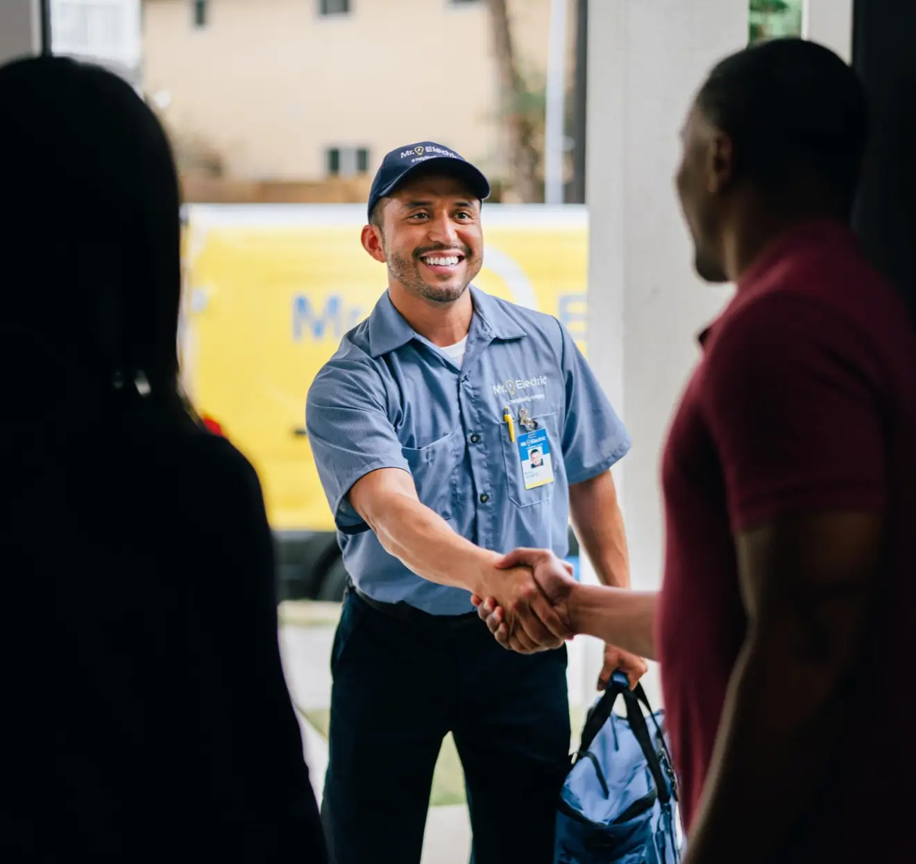 Smiling electrician in a blue uniform shirt and cap, standing at the entrance of a home, shaking hands with a client while holding a blue tool bag, with a yellow service van visible in the background and another person in silhouette in the foreground.