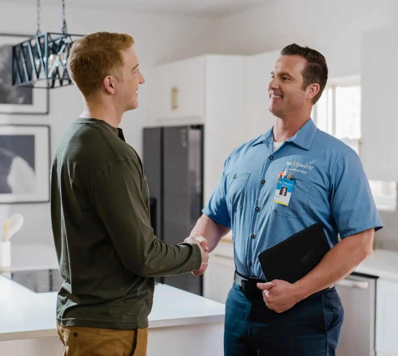 Smiling electrician in a blue uniform shaking hands with a customer in a modern kitchen, holding a black tablet portfolio, under a decorative hanging light fixture.