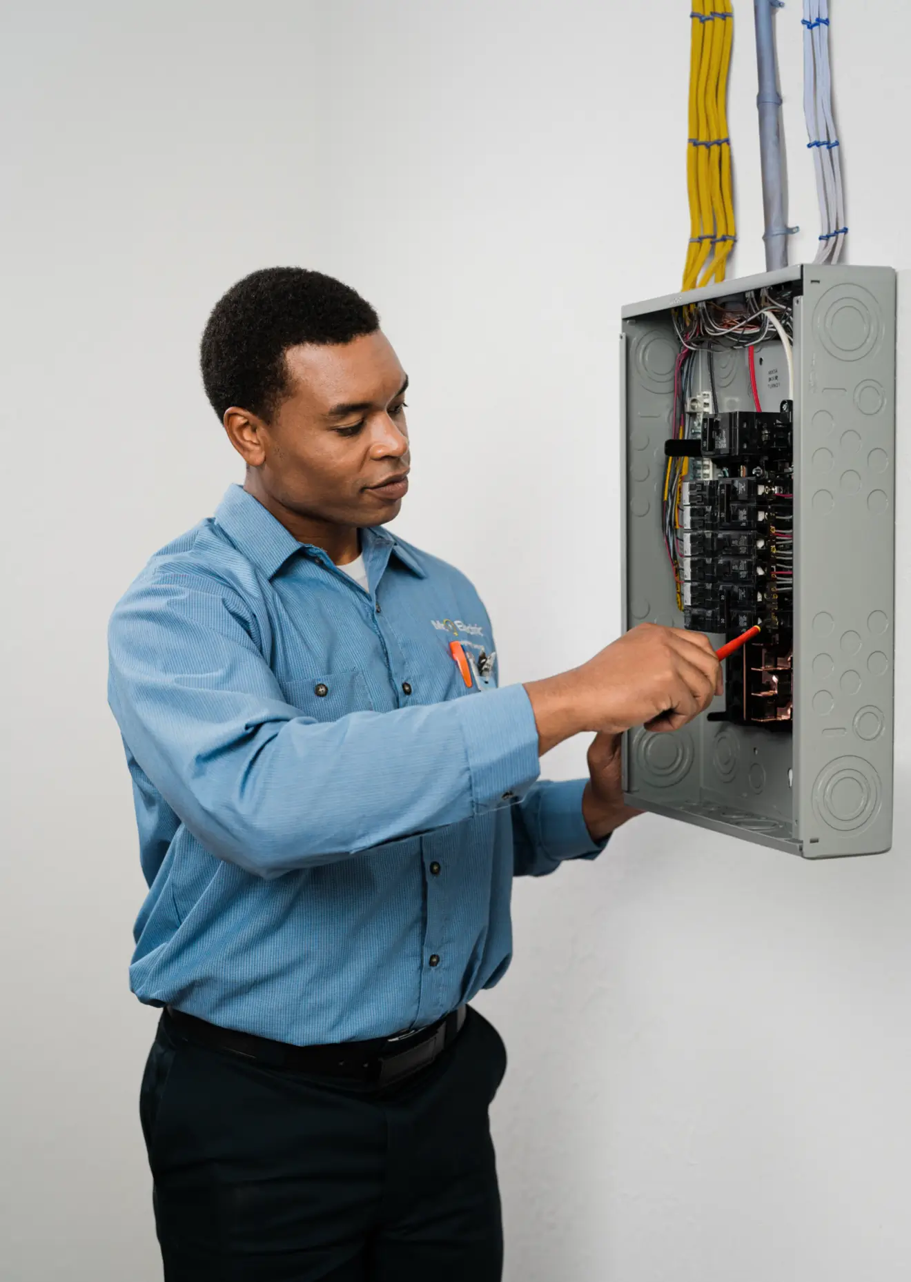 Electrician in blue uniform inspecting and working on an open electrical breaker panel attached to a wall.