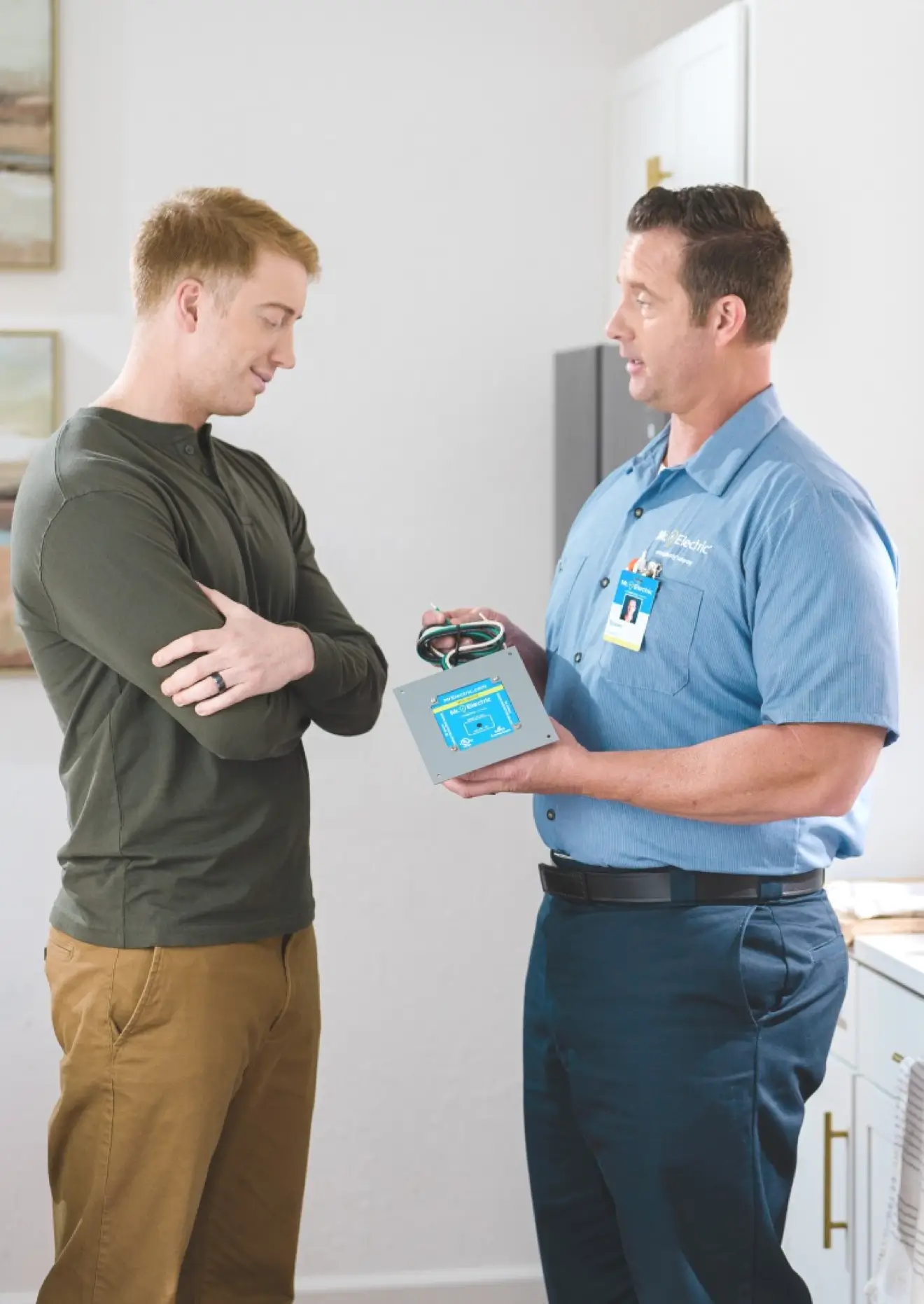 Electrician in blue shirt holding a whole home surge protector device while explaining installation details to a homeowner dressed in an olive green shirt, standing together in a modern home interior.
