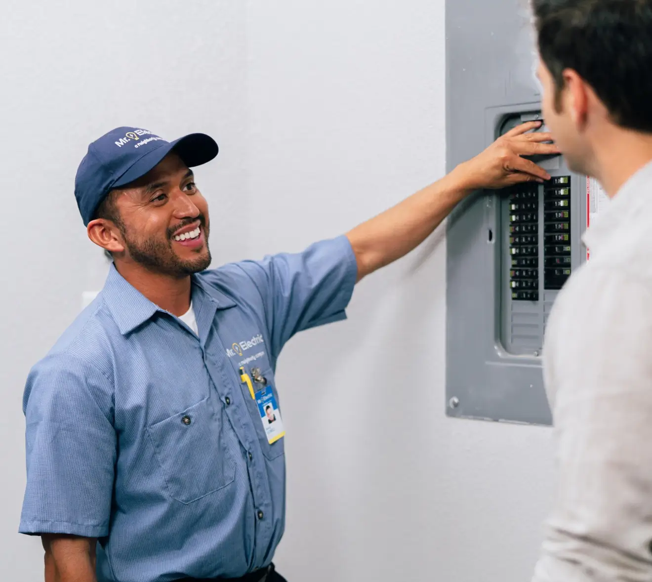 Friendly electrician in blue uniform and cap smiling while standing beside an open circuit breaker panel, talking to a customer.
