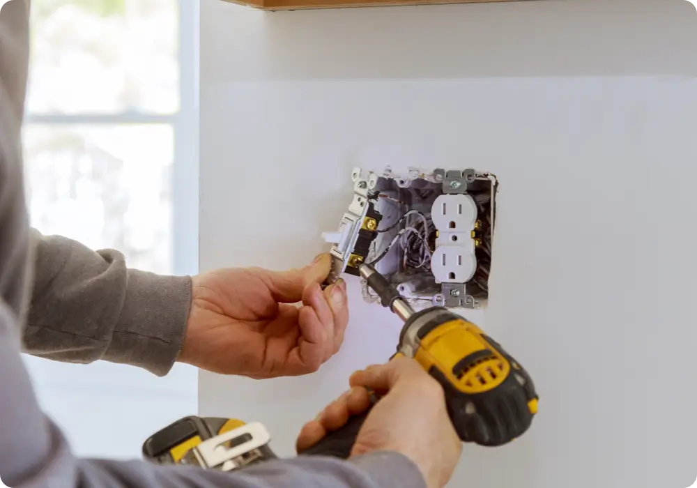 An electrician is seen from the shoulders down, wearing a grey long-sleeved shirt, using a yellow and black power drill to work on the wiring of a white electrical outlet in a white wall.