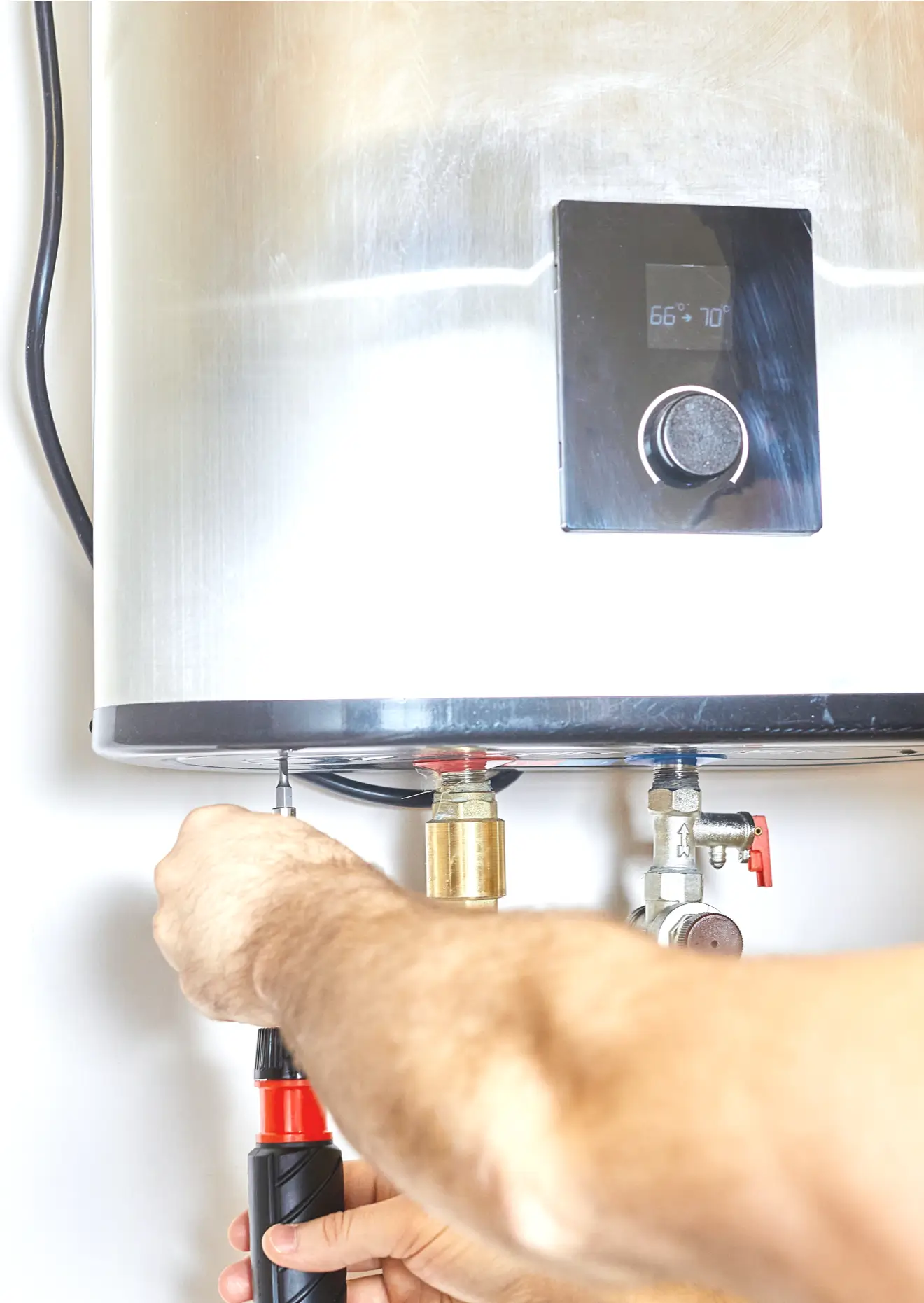 Hands of an installer using a screwdriver on the bottom of a brushed stainless steel tankless water heater mounted on a white wall; temperature display and water pipes visible at the base.