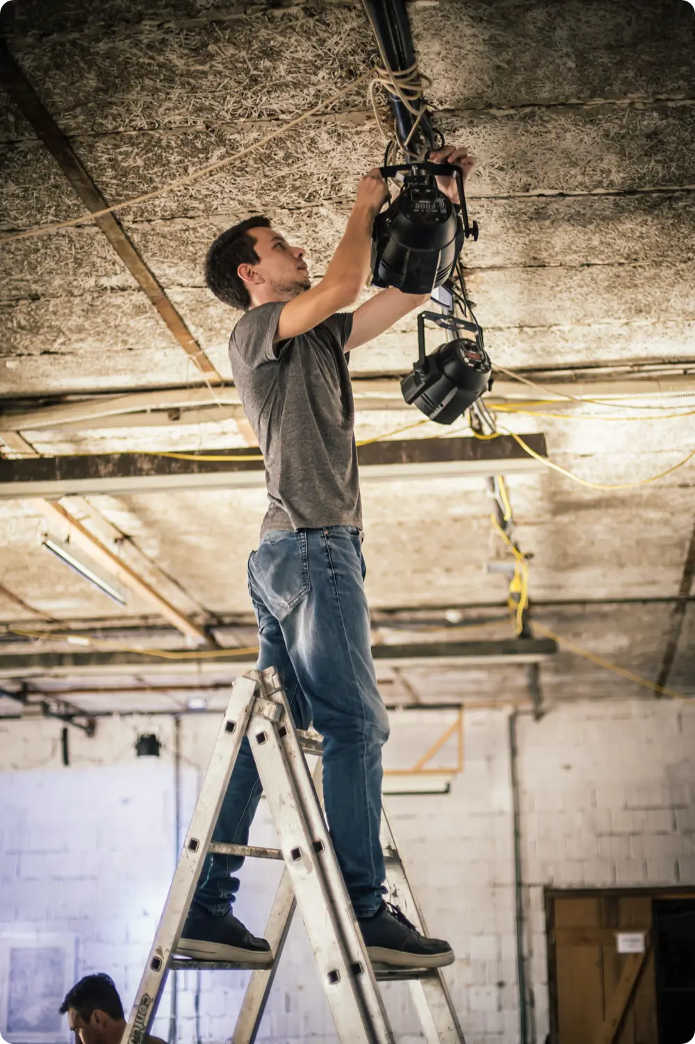 A young man wearing a gray T-shirt and blue jeans is standing on a silver A-frame ladder, reaching up to adjust black theater spotlight fixtures hanging from a ceiling with exposed structural elements and wiring.