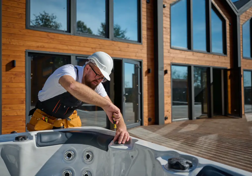 Bearded technician with white safety helmet and black overalls installing an outdoor hot tub on a wood patio in front of large windows under clear skies.