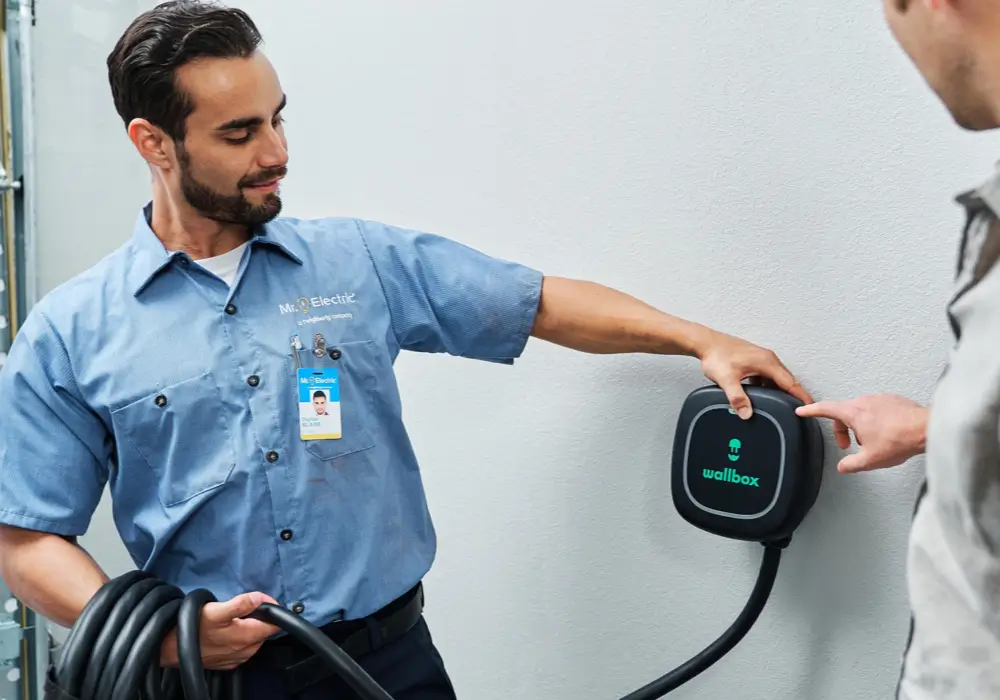 Male service professional in a blue work shirt and black pants demonstrating a Wallbox electric vehicle charger on an exterior wall, holding the cable and pointing to the charger panel for a customer.
