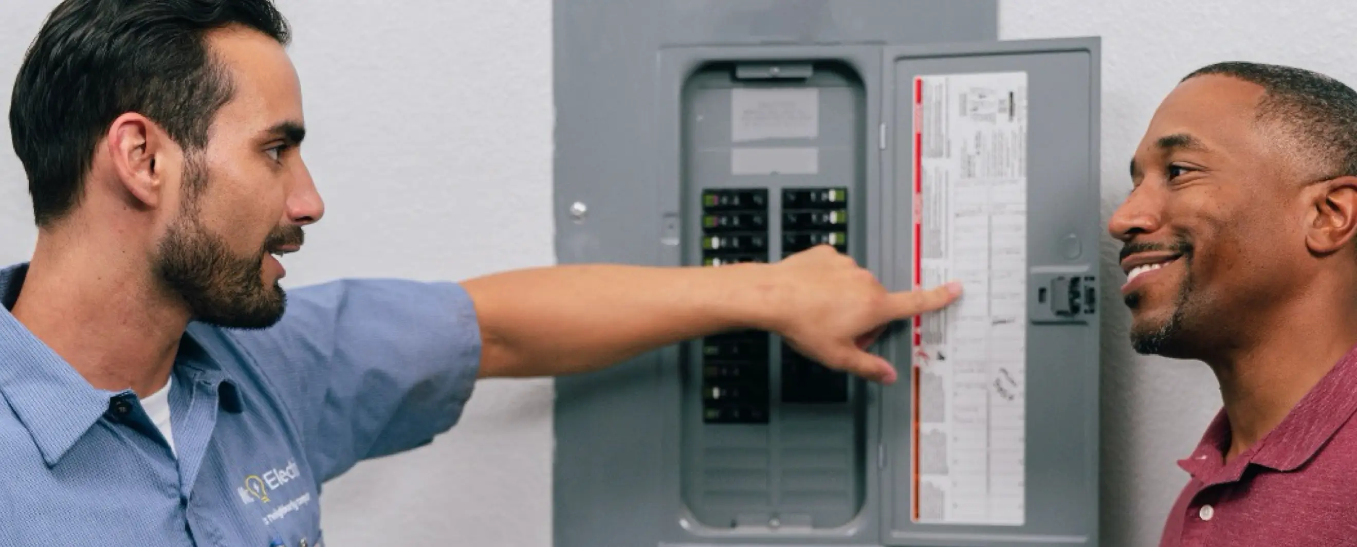 Service professional in blue uniform pointing and explaining features of a circuit breaker panel to a smiling customer.