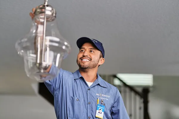 A smiling Mr. Electric electrician reaches for a pendant light fixture suspended from a ceiling.